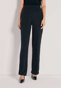 Vrouw in zwarte hoge taille, rechte, tekstuur broek en zwarte puntige hakken, staand tegen een eenvoudige lichte muur.