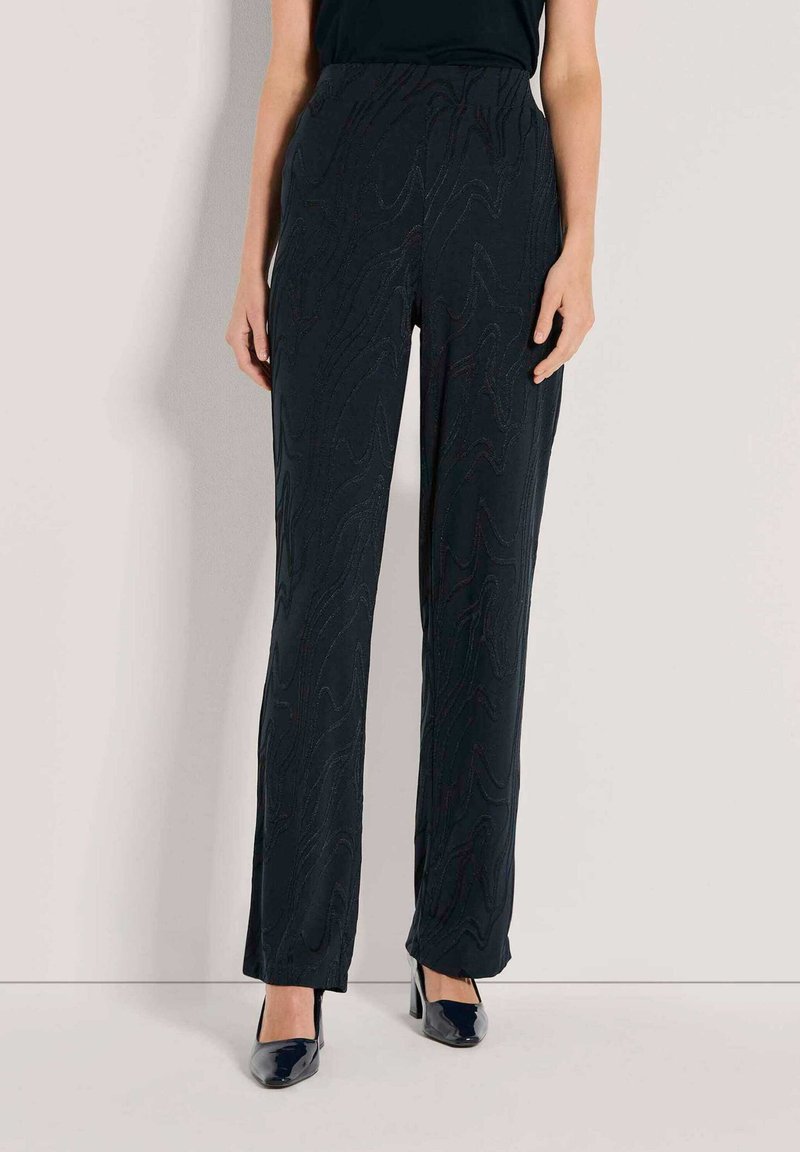 Vrouw in zwarte hoge taille, rechte, tekstuur broek en zwarte puntige hakken, staand tegen een eenvoudige lichte muur.