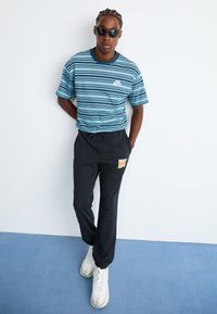 Mann mit schwarzen Sonnenbrillen, blau gestreiftem Nike-T-Shirt, schwarzen Hosen mit einem Patch und weißen Sneakers, der vor einer weißen Wand steht.