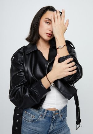 Mujer con cabello oscuro a la altura de los hombros, vestida con una chaqueta de cuero negra, camiseta blanca, vaqueros azules y dos pulseras plateadas enlazadas sobre cordones marrones.