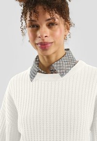 Biały, dziergany sweter o fakturowanej strukturze z okrągłym dekoltem, nałożony na szarą koszulę w kratę. Modelka ma kręcone włosy i srebrne kolczyki-koła.