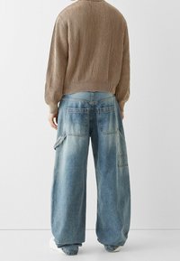 Světle modré džíny s širokými nohavicemi a vybledlým efektem, s dvěma zadními kapsami a nákladní kapsou na levé straně. Nošeny s žebrovaným hnědým svetrem.