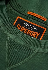 Grüner Baumwollpullover mit geripptem Kragen. Verfügt über ein schwarzes Etikett mit orangener Schrift, auf dem "Qualität" und "Superdry" steht, sowie ein oranges Label.