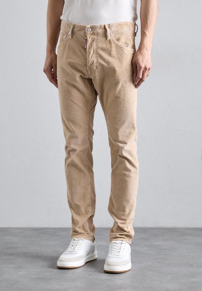 Un homme portant un pantalon en velours côtelé beige et des baskets blanches se tient sur un sol gris contre un mur léger gris uni.