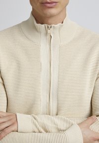 Beige zip-up trui met een gestructureerd ribbelpatroon, hoge kraag en aansluitende manchetten, gemaakt van een zachte, rekbare stof.