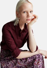 Burgundy button-up shirt with rolled sleeves, paired with a patterned purple skirt featuring floral designs in white and pink. Silver hoop earrings.