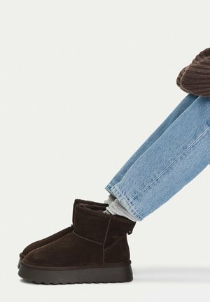 Personne portant un jean bleu clair, des chaussettes grises et des bottines en daim marron foncé avec des semelles noires, assise sur un fond blanc.