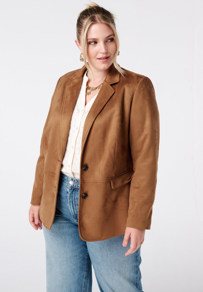 Blazer en suédine marron avec revers en pointe, deux boutons et poches plaquées, porté sur un haut crème et un jean bleu clair.