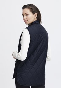 Mornarska jakna bez rukava s visokim ovratnikom, u quilted stilu, s bočnim prorezima. Nošena preko bijele majice s dugim rukavima, prikazujući glatku teksturu tkanine.