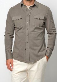 Chemise grise à manches longues en coton doux, avec deux poches poitrine à rabat et fermeture pression, et un ourlet arrondi.