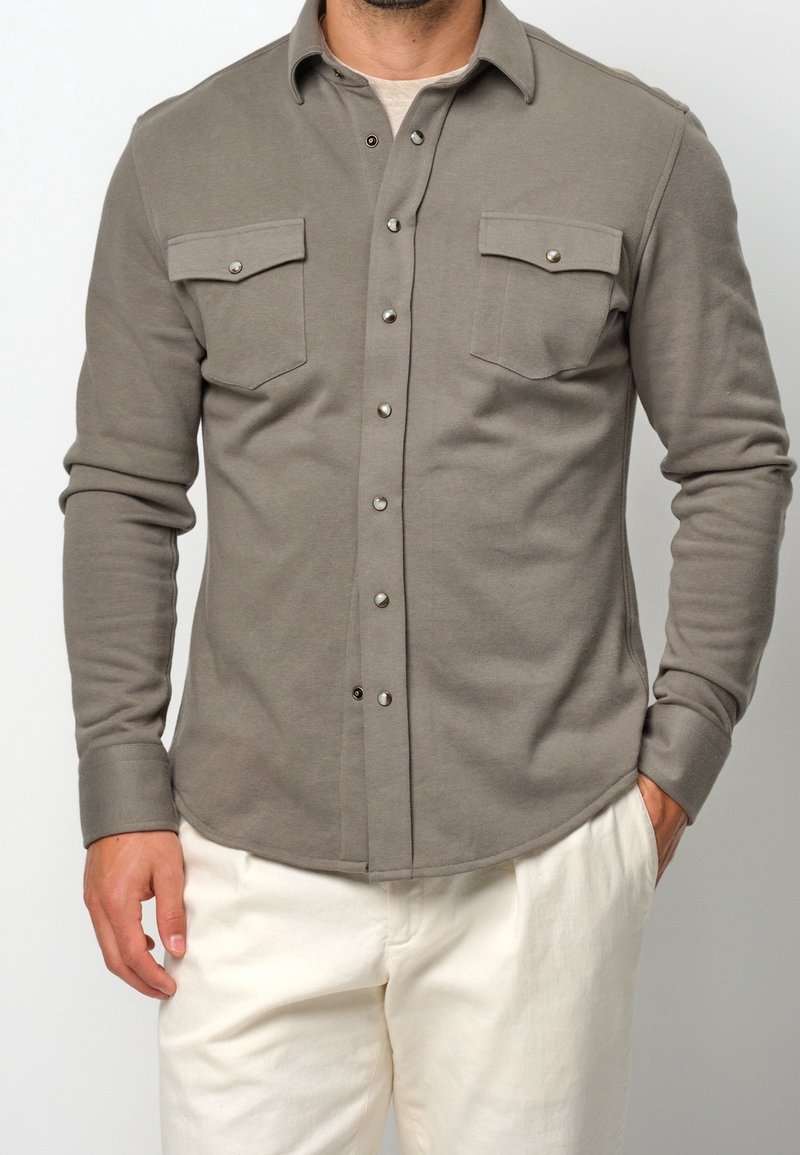 Chemise grise à manches longues en coton doux, avec deux poches poitrine à rabat et fermeture pression, et un ourlet arrondi.