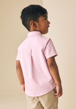 Jeune garçon aux cheveux courts noirs portant une chemise rose clair à manches courtes et un pantalon beige, debout et regardant vers le haut à droite.