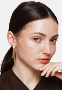 Gouden geometrische oorbellen hebben meerdere verticale strengen en tonen een strak ontwerp met een gladde textuur. Het model draagt een donkerbruine top.