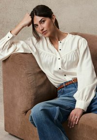 Witte katoenen blouse met een knoopsluiting, gecombineerd met hooggemeten blauwe jeans en een gevlochten bruine riem, zittend op een gestructureerde brown bank.