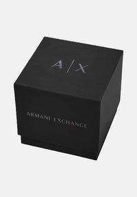 Caja negra de Armani Exchange con el logo plateado A|X en la parte superior y el texto de Armani Exchange en el lado frontal, aislada sobre un fondo claro.