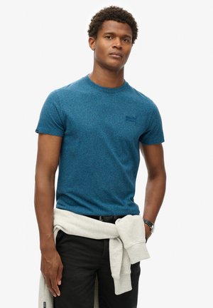 Blauwe heide korte mouwen t-shirt met ronde hals en subtiele logo; gecombineerd met zwarte broek en een lichtgrijze hoodie die om de taille is geknoopt.