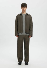 Brūna rāvējslēdzēja jaka pār pelēku džemperi, komplektā ar brūnām biksēm. Audums ir gluds, ar piegriezumu, kas ir gan stilīgs, gan ērtums. Melni apavi.