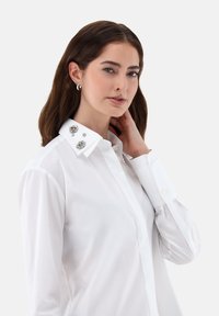 Camicia bianca con colletto rifinito, caratterizzato da decorazioni metalliche. Maniche lunghe con polsini abbottonati; texture liscia.