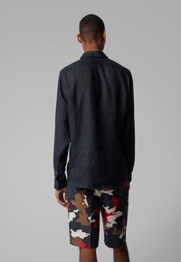 Muž otočený zády, oblečený v tmavé košili s dlouhým rukávem a šortkách s kamuflážním vzorem v červených, hnědých, bílých a černých tónech.