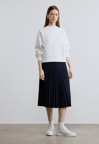 Sweat-shirt blanc en coton avec poignets côtelés, associé à une jupe plissée bleu marine, porté avec des baskets blanches et des socquettes, debout sur un sol neutre.