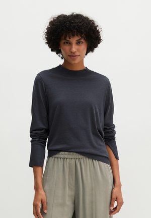 Vrouw met krullend haar, gekleed in een donkergrijze top met lange mouwen en losse lichtgrijze broek, staande tegen een effen achtergrond.