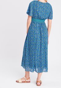 Robe longue à motif bleu avec manches courtes, taille smockée et tissu fluide. Associée à des sandales marron clair avec une lanière tressée.