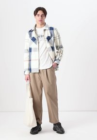 Jackete în carouri alb, albastru și bej peste un tricou alb, asortat cu pantaloni bej și pantofi negri. Ținând o geantă de umăr bej.