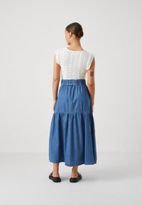 Denim maxi krilo z nabranim pasom in večstopenjsko zasnovo, v kombinaciji z belo bluzo z odprtimi očesi, ki ima kratke rokave in vertikalne stripe.