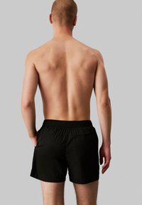 Pantaloni da nuoto neri con vita elastica, in tessuto leggero e dal taglio rilassato, mostrati da dietro. Senza motivi o dettagli visibili.