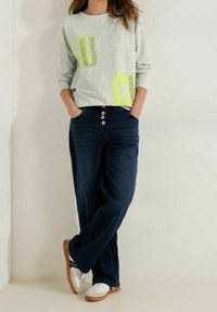 Sudadera gris claro con letras en amarillo neón, combinada con jeans azul oscuro con cierre de cinco botones y zapatillas claras con detalles en negro.