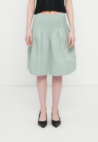 Φούστα μήκους μέχρι τα γόνατα σε ανοιχτό πράσινο χρώμα με πιέτες, λεία υφή και φαρδιά μέση. Συνδυάζεται με μαύρα παπούτσια μυτερά, διακοσμημένα με κόμπο.