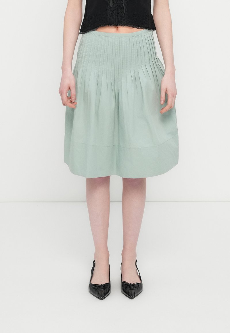 Φούστα μήκους μέχρι τα γόνατα σε ανοιχτό πράσινο χρώμα με πιέτες, λεία υφή και φαρδιά μέση. Συνδυάζεται με μαύρα παπούτσια μυτερά, διακοσμημένα με κόμπο.