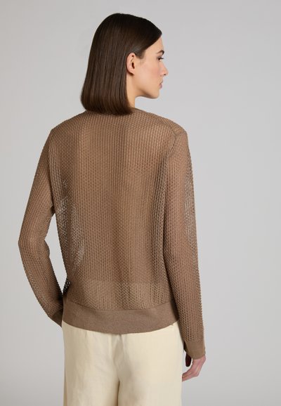 Vrouw met steil donker haar, gekleed in een doorschijnende bruine, lange mouwen gebreide trui en lichtgekleurde broek, zijwaarts kijkend tegen een effen achtergrond.