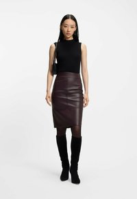 Gonna a tubino in pelle bordeaux abbinata a un top senza maniche nero. Modella che indossa stivali neri in suede fino al ginocchio, tenendo una piccola borsa nera.