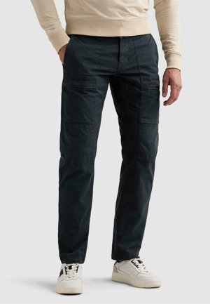 Homme portant un pantalon cargo foncé avec des poches latérales et des baskets blanches, debout avec une main dans la poche sur un fond uni.