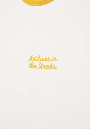 Bijela tkanina sa žutim ovratnikom i žutim izvezenim tekstom "Art lives in the Streets" centriranim ispod ovratnika.
