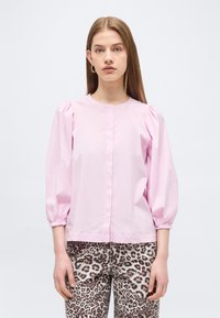 Jeune femme aux cheveux raides portant un chemisier rose clair à manches bouffantes et un pantalon imprimé léopard sur un fond uni.