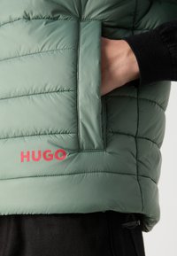 Jachetă căptușită în verde stins, cu o textură în dungi, buzunar lateral și un logo roșu „HUGO” proeminent la tiv. Detalii negre la mansete vizibile.