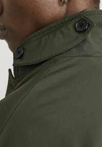 Giacca verde scuro con colletto a punta, dettagli con bottoni e texture di tessuto liscia. Focus sui dettagli del colletto e sulla cucitura della spalla.