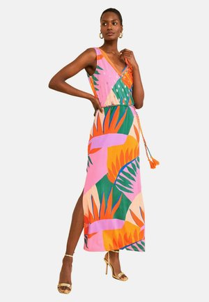 Frau trägt ein ärmelloses, farbenfrohes Maxikleid mit geometrischen tropischen Blattmustern, kombiniert mit goldenen High-Heel Sandalen und Creolen.