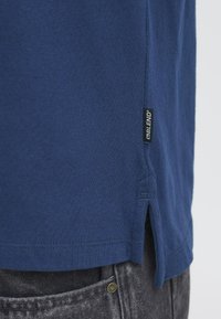Marineblå bomulds t-shirt med sideslids, med et synligt sort label på den nederste søm; sat sammen med grå denimbukser.