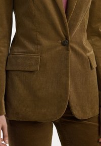 Hnědý kordový blejzr s vertikálním žebrováním, s jedním knoflíkem a dvěma předními kapsami. Textura se zdá být měkká; přiléhavý design.