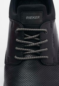 Black RIEKER shoe with a textured leather upper, grey laces, and perforations. The brand logo is embossed on a black patch.