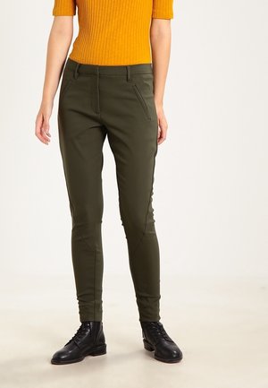Femme portant un pantalon slim vert olive, des bottines noires à lacets et un haut à manches courtes côtelé couleur moutarde, debout sur un fond neutre.