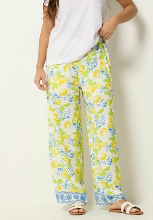 Kobieta nosząca spodnie z szerokimi nogawkami z żółtym nadrukiem cytryn i zielonych liści, w połączeniu z białymi sandałami i białą koszulką.