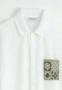 Chemise blanche à manches courtes avec un tissu texturé, dotée d'une poche plaquée ornée de motifs brodés verts et d'un col.