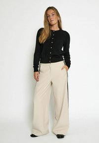 Svart stickad cardigan med rund halsringning och knäppning, kombinerad med beige vida byxor med sidostripes och framfickor.
