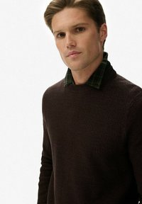 Bruine gestructureerde gebreide trui over een groen geruit colshirt. De trui heeft een ronde halslijn en getailleerde mouwen, met een subtiel patroon.