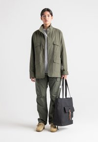 Veste olive avec poches utilitaires, associée à un pantalon cargo assorti et des chaussures beiges. Tient un sac fourre-tout noir avec une sangle en cuir.