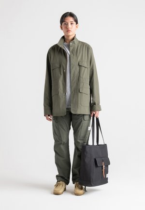 Olivovo zelená úžitková bunda s dvoma náprsními vreckami, v kombinácii s farebnými cargo nohavicami a čiernou taškou na rameno s klopou a koženým popruhom.